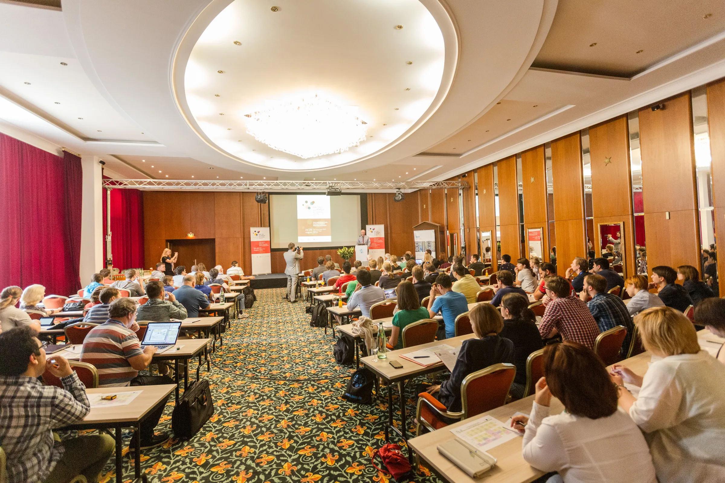 Eine gut besuchte Konferenz mit Vortrag, Projektion und Publikum in einem großen Saal.