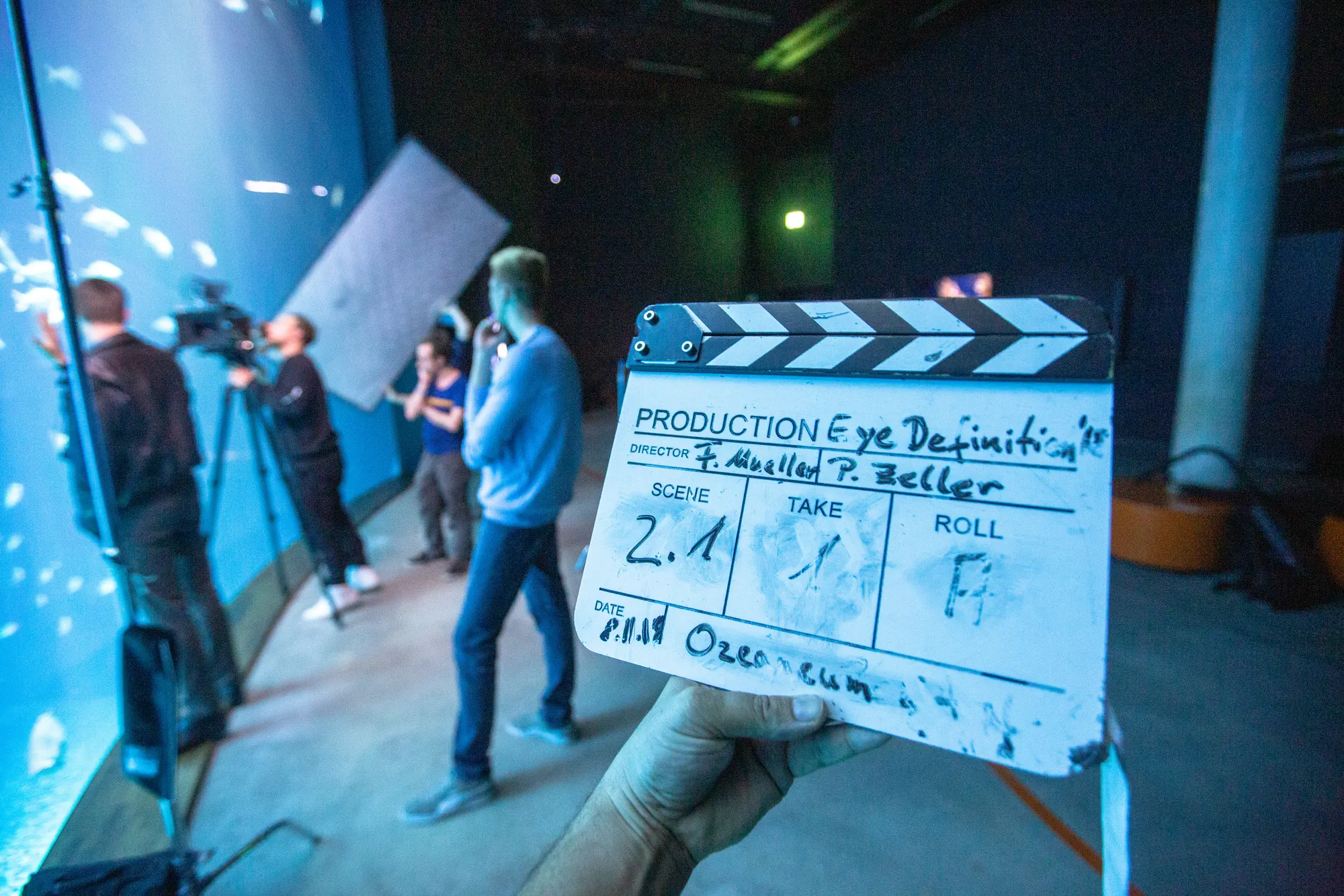 Eine Filmklappe vor einem laufenden Videodreh mit Kamera, Licht und Team.