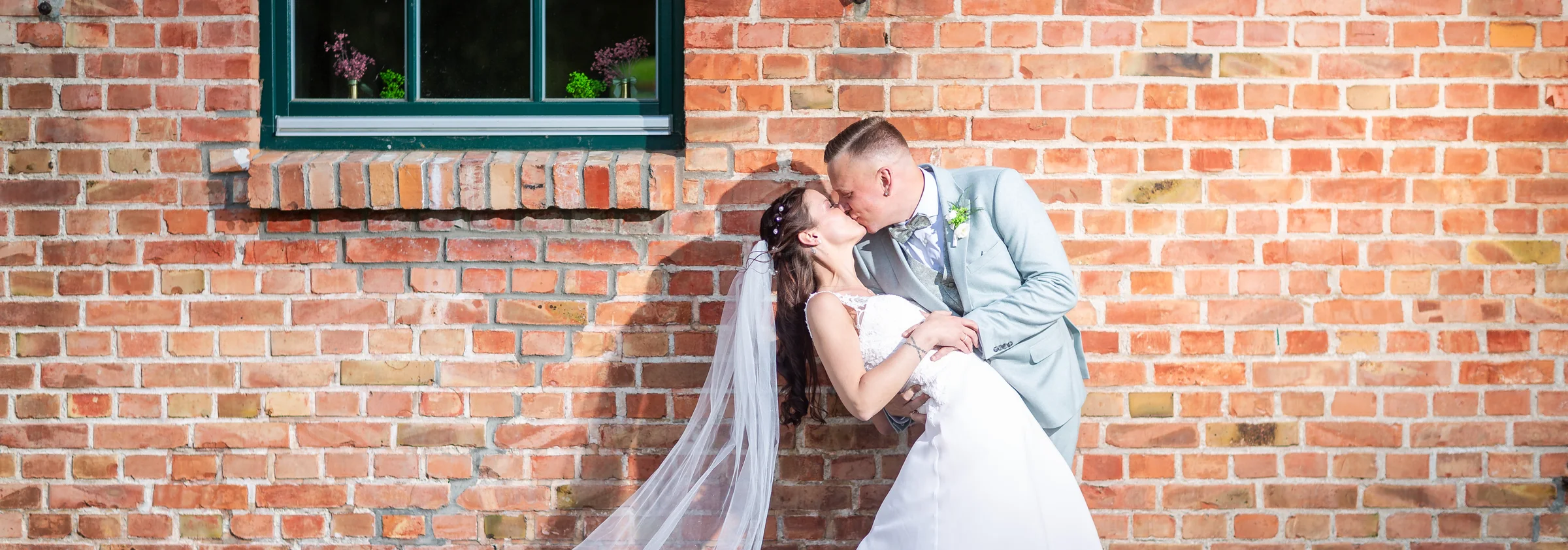 Ein Hochzeitspaar küsst sich vor einer sonnenbeschienenen Backsteinwand.