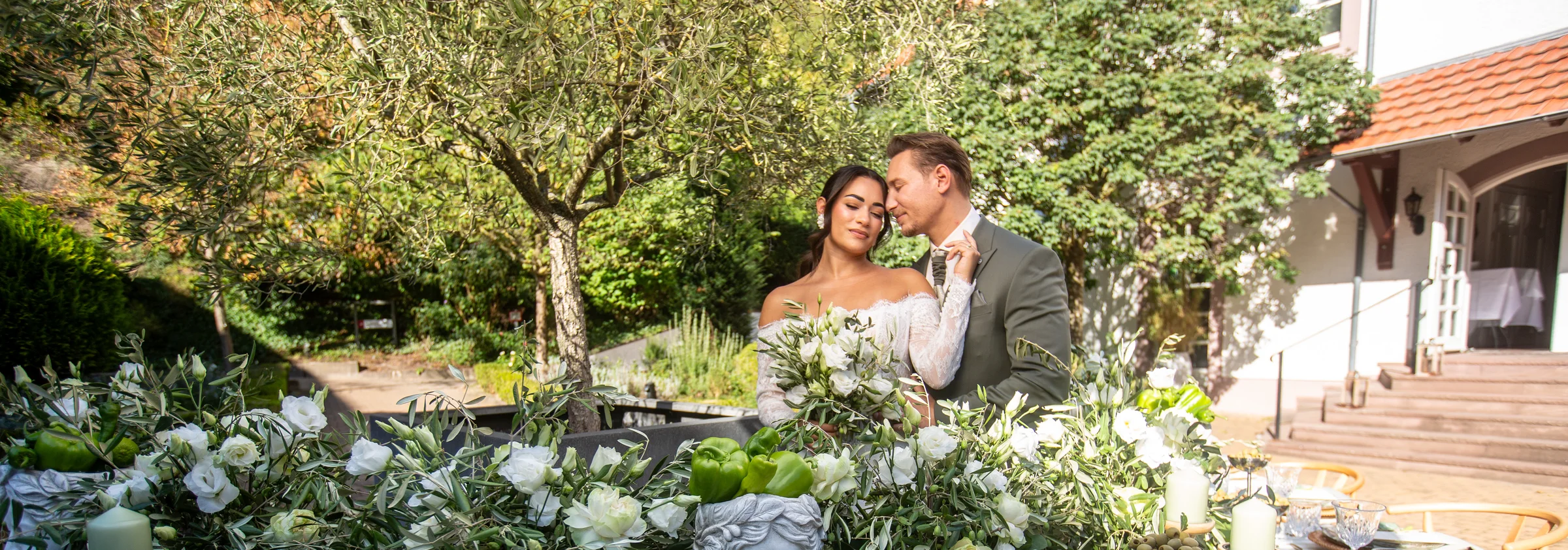 Ein Hochzeitspaar steht eng beieinander hinter einer mediterran gedeckten Festtafel.